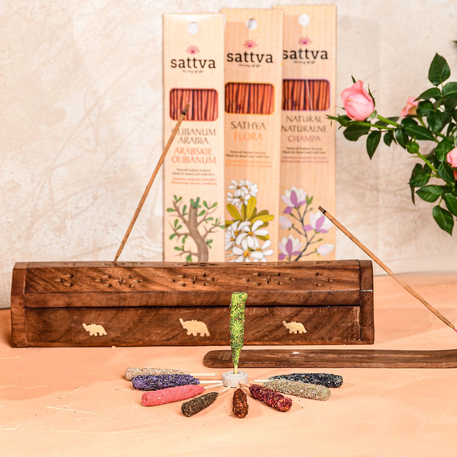 Naturalne kadzidełka Sattva Ayurveda – zestaw aromatycznych pałeczek i stożków w drewnianej szkatułce z motywem słoni. W tle opakowania kadzidełek o zapachu arabskiego olibanum, champa i flora, obok świeże kwiaty róż