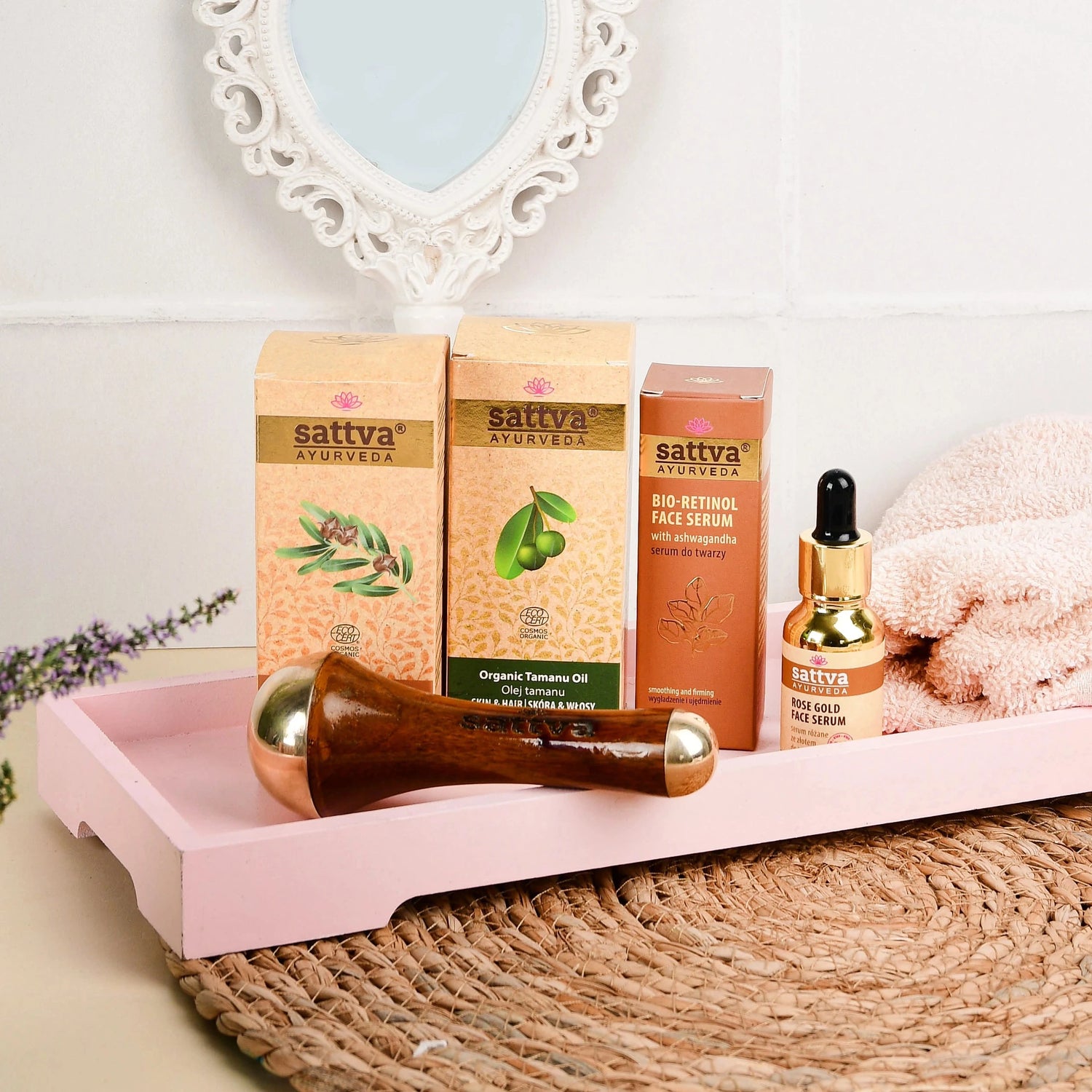 Naturalne kosmetyki Sattva Ayurveda do pielęgnacji twarzy – Bio Retinol Face Serum z ashwagandhą, Organic Tamanu Oil oraz Rose Gold Face Serum, zaprezentowane na różowej tacy z drewnianym masażerem Kansa wand, ręcznikiem i lusterkiem w kształcie serca – przytulna aranżacja w łazienkowym stylu